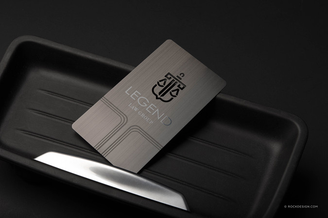 Black brushed metal business card with cutout logo and silver etched text for Legend Law Group, displayed on a sleek black tray.